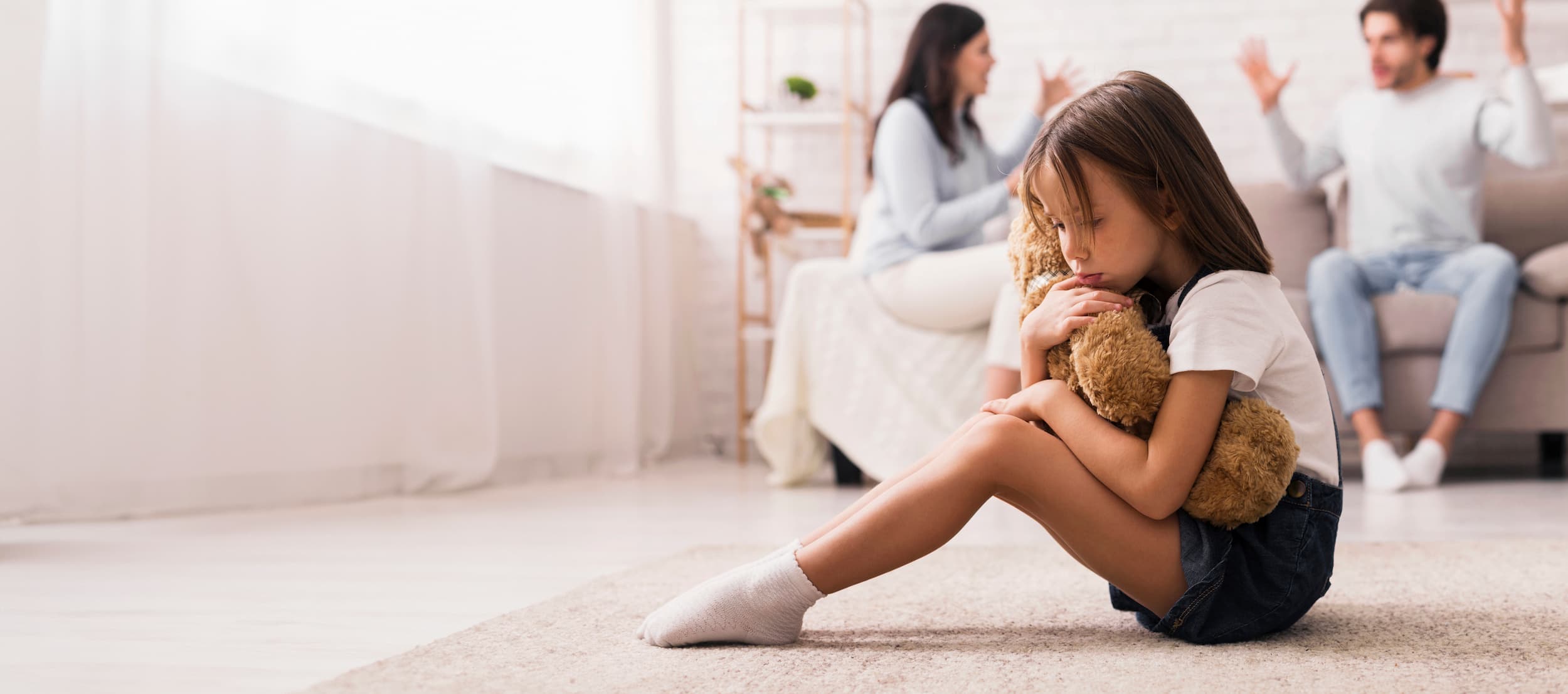 Upset young girl hugging teddy bear, parents arguing in the background.